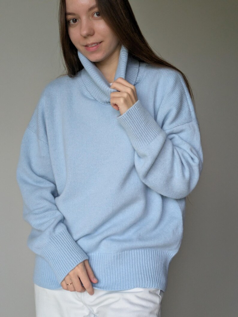 May include: A light blue sweater with a turtleneck collar and ribbed cuffs. The sweater has a relaxed fit and is made of a soft material. The model is wearing white pants. This is a casual, everyday sweater.
