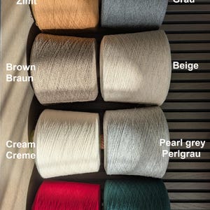 Op de afbeelding: Acht cilindervormige garenspoelen gerangschikt in twee kolommen. De kleuren zijn onder andere kaneel, grijs, bruin, beige, cr&egrave;me, parelgrijs, rood en groen. Tekstlabels naast elke spoel identificeren de kleuren in het Engels en Duits, zoals Cinnamon Zimt.