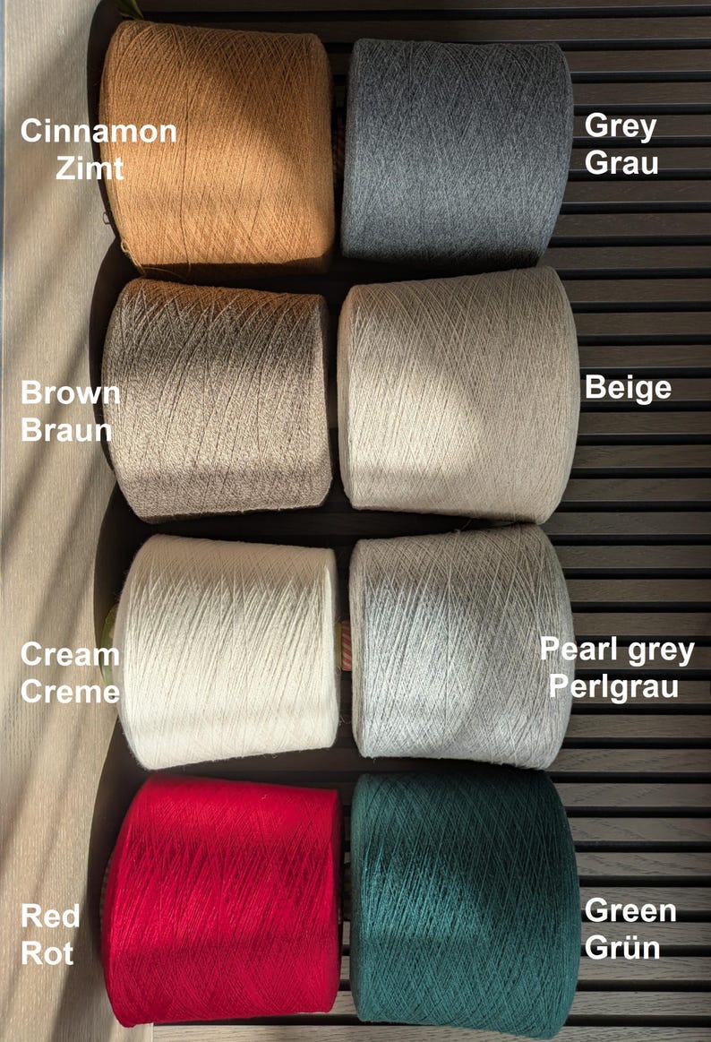 Op de afbeelding: Acht cilindervormige garenspoelen gerangschikt in twee kolommen. De kleuren zijn onder andere kaneel, grijs, bruin, beige, cr&egrave;me, parelgrijs, rood en groen. Tekstlabels naast elke spoel identificeren de kleuren in het Engels en Duits, zoals Cinnamon Zimt.