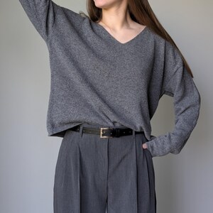 Puede incluir: Un su&eacute;ter gris de cuello en V con mangas largas y un cintur&oacute;n negro usado con pantalones grises plisados.
