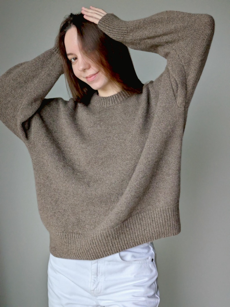 May include: A long-sleeved, brown knit sweater featuring a crew neckline and ribbed cuffs and hem, paired with white denim jeans. The sweater has a loose, relaxed fit with a textured, heathered appearance throughout the fabric.