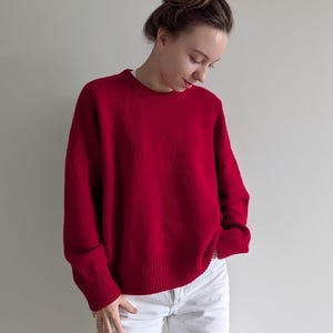May include: A vibrant red long-sleeved sweater with a crew neckline and ribbed cuffs and hem. The sweater features a relaxed fit and is paired with plain white pants. The knit fabric has a soft texture and a solid, deep red color throughout.