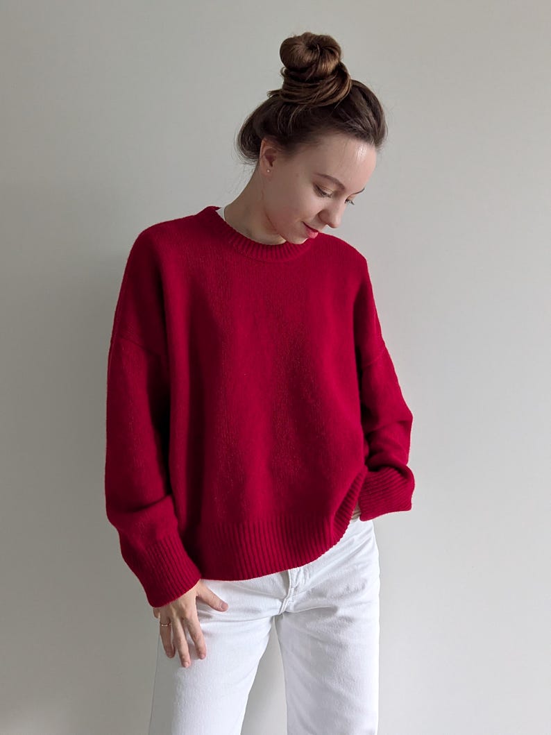 May include: A vibrant red long-sleeved sweater with a crew neckline and ribbed cuffs and hem. The sweater features a relaxed fit and is paired with plain white pants. The knit fabric has a soft texture and a solid, deep red color throughout.