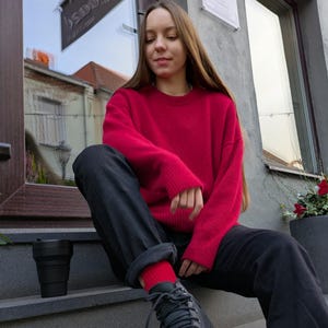 May include: A bright red long-sleeved crewneck sweater paired with black trousers, red socks, and black lace-up sneakers. A black collapsible cup sits on the step nearby. The outfit features a vibrant knit texture and a casual, relaxed fit.