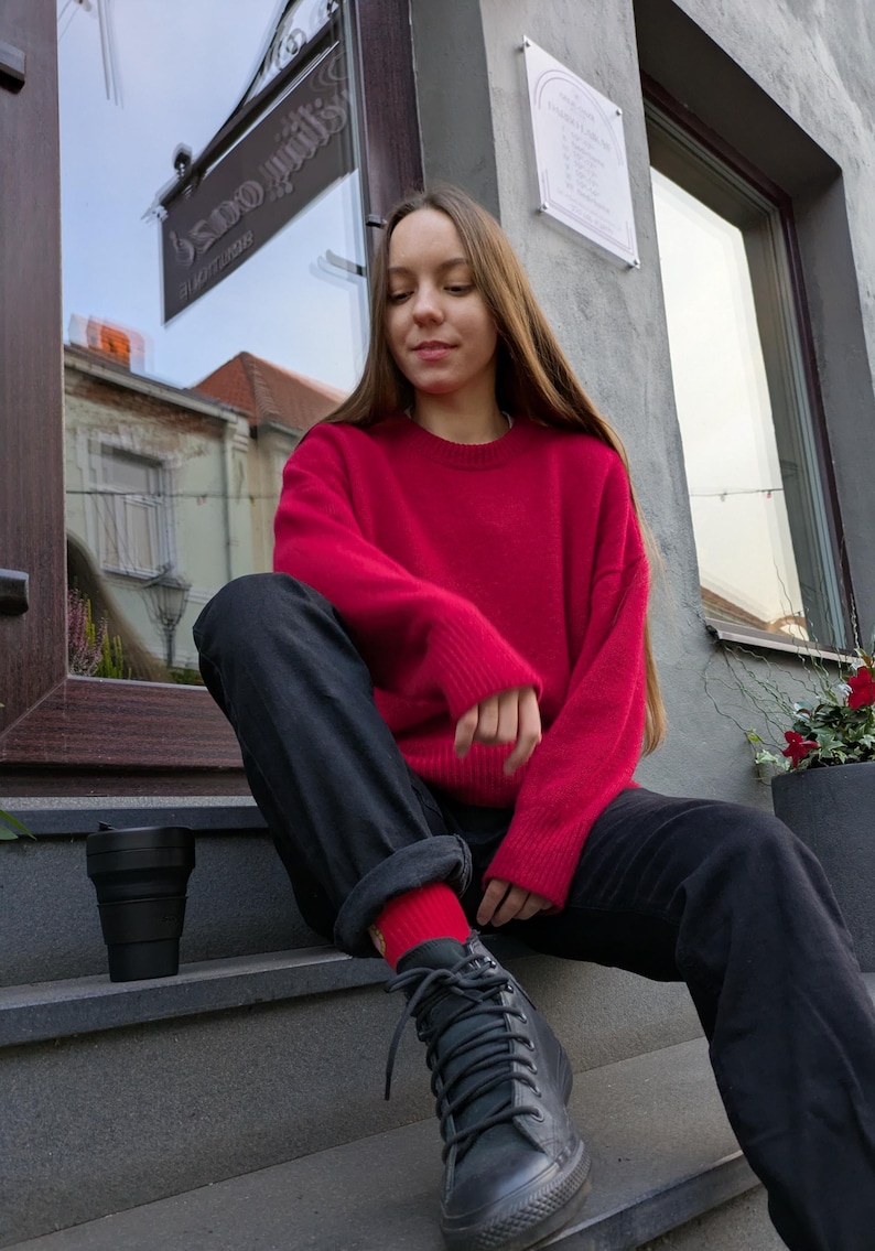 May include: A bright red long-sleeved crewneck sweater paired with black trousers, red socks, and black lace-up sneakers. A black collapsible cup sits on the step nearby. The outfit features a vibrant knit texture and a casual, relaxed fit.