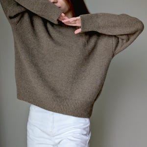 May include: A long-sleeved, brown knit sweater featuring a crew neckline and ribbed cuffs and hem, paired with solid white pants. The sweater has a relaxed fit with dropped shoulders and a textured knit pattern throughout the garment.