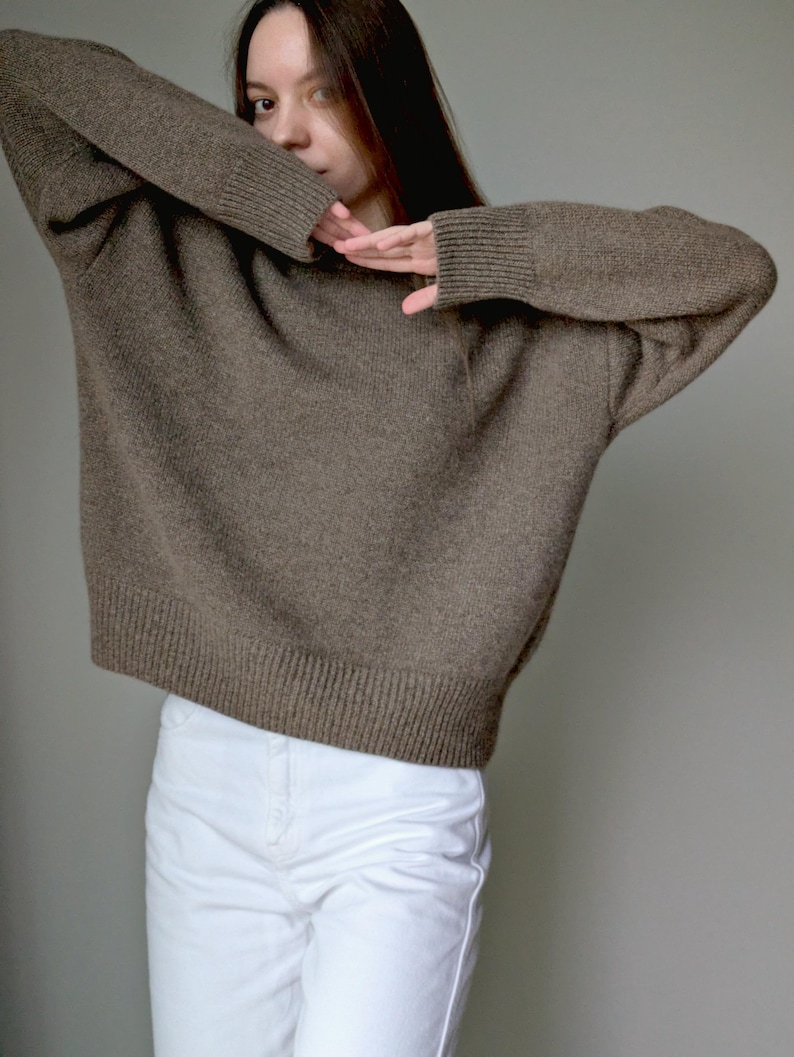 May include: A long-sleeved, brown knit sweater featuring a crew neckline and ribbed cuffs and hem, paired with solid white pants. The sweater has a relaxed fit with dropped shoulders and a textured knit pattern throughout the garment.