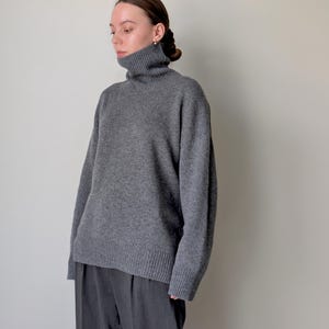 Op de afbeelding: Een antracietgrijze trui met lange mouwen, een hoge col, verlaagde schouders en geribbelde gebreide details bij de manchetten en de zoom, gecombineerd met een donkergrijze broek met een zichtbare verticale vouw aan de voorzijde.