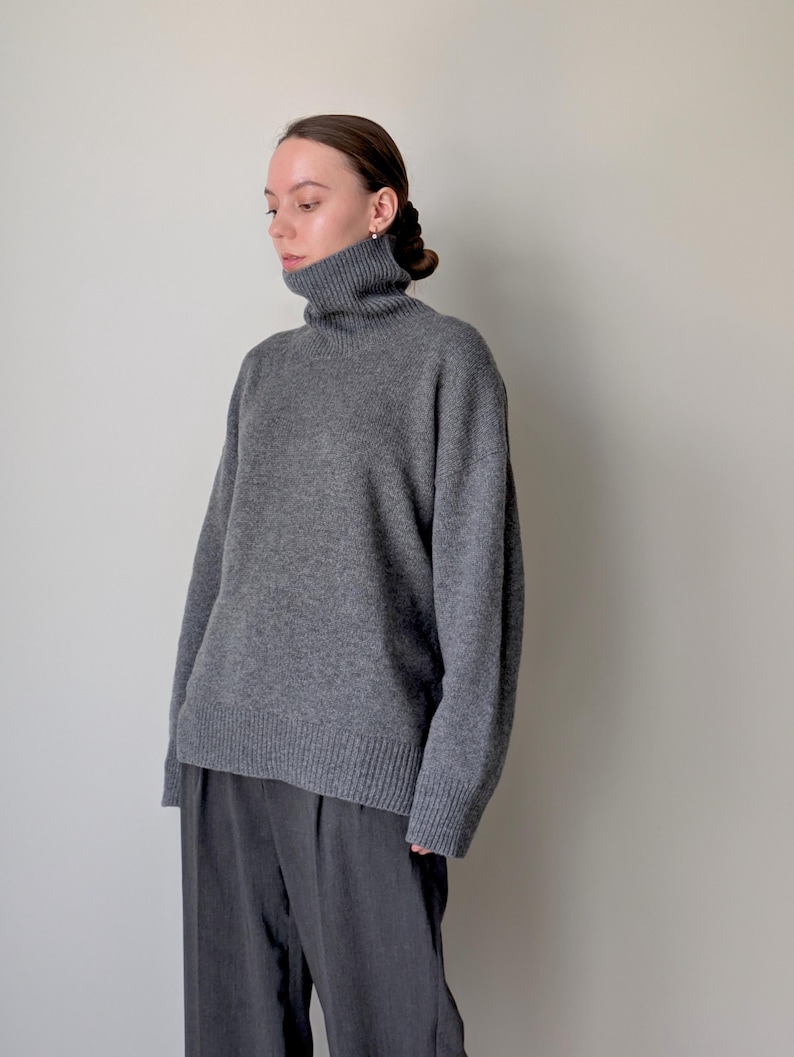Op de afbeelding: Een antracietgrijze trui met lange mouwen, een hoge col, verlaagde schouders en geribbelde gebreide details bij de manchetten en de zoom, gecombineerd met een donkergrijze broek met een zichtbare verticale vouw aan de voorzijde.