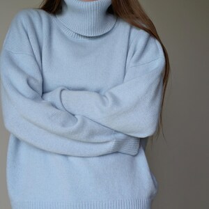 May include: A light blue turtleneck sweater. The sweater has long sleeves and a ribbed hem. The model is wearing the sweater with their arms crossed. The sweater is made of a soft, knitted material.