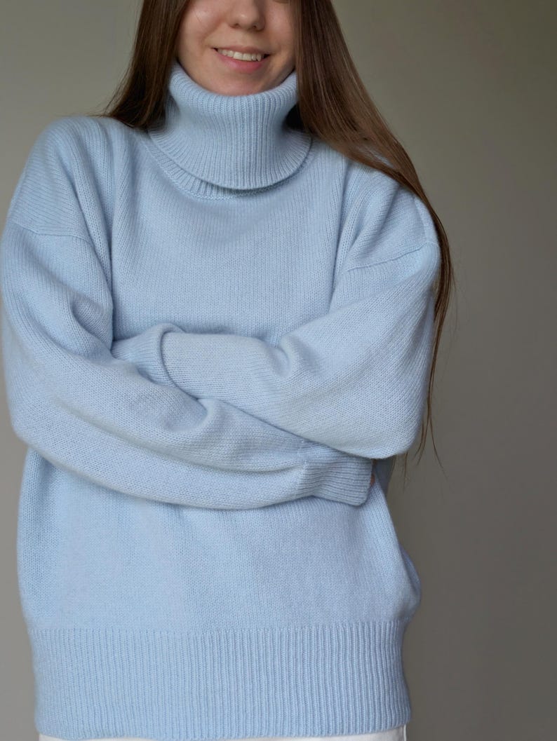 May include: A light blue turtleneck sweater. The sweater has long sleeves and a ribbed hem. The model is wearing the sweater with their arms crossed. The sweater is made of a soft, knitted material.