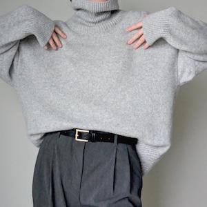 Op de afbeelding: Een lichtgrijze oversized coltrui met lange mouwen en geribbelde boorden, gecombineerd met een donkergrijze pantalon met plooien en een zwarte leren riem met een goudkleurige rechthoekige gesp, gedragen als een complete outfit.