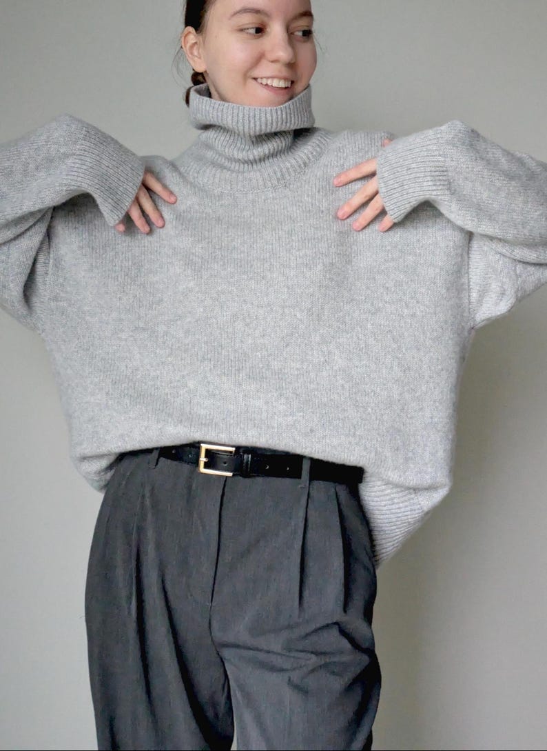 Op de afbeelding: Een lichtgrijze oversized coltrui met lange mouwen en geribbelde boorden, gecombineerd met een donkergrijze pantalon met plooien en een zwarte leren riem met een goudkleurige rechthoekige gesp, gedragen als een complete outfit.