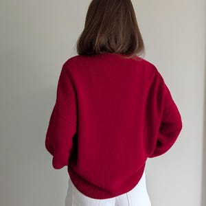 May include: A long-sleeved, deep red knit sweater featuring a ribbed crew neckline, ribbed cuffs, and a ribbed hem. The sweater is paired with white denim pants that have visible back pockets and a structured waistband, creating a simple contrast.