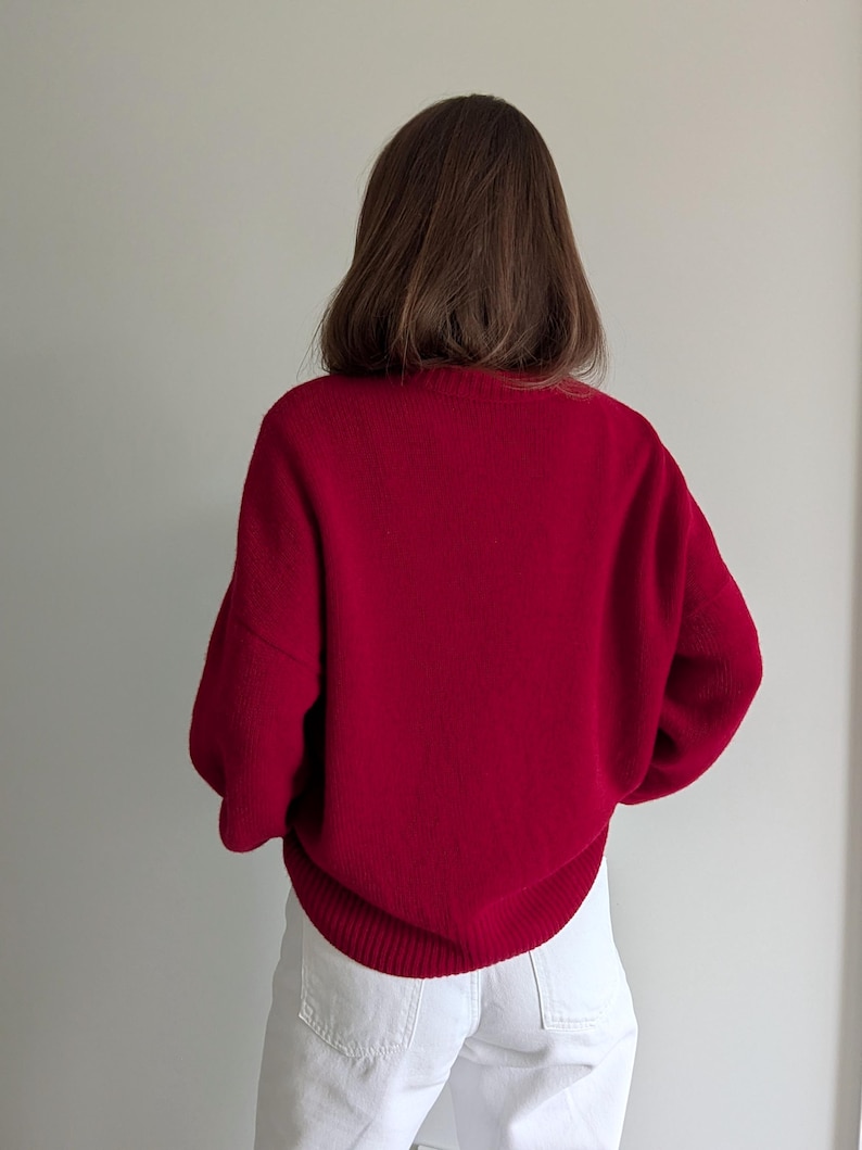 May include: A long-sleeved, deep red knit sweater featuring a ribbed crew neckline, ribbed cuffs, and a ribbed hem. The sweater is paired with white denim pants that have visible back pockets and a structured waistband, creating a simple contrast.