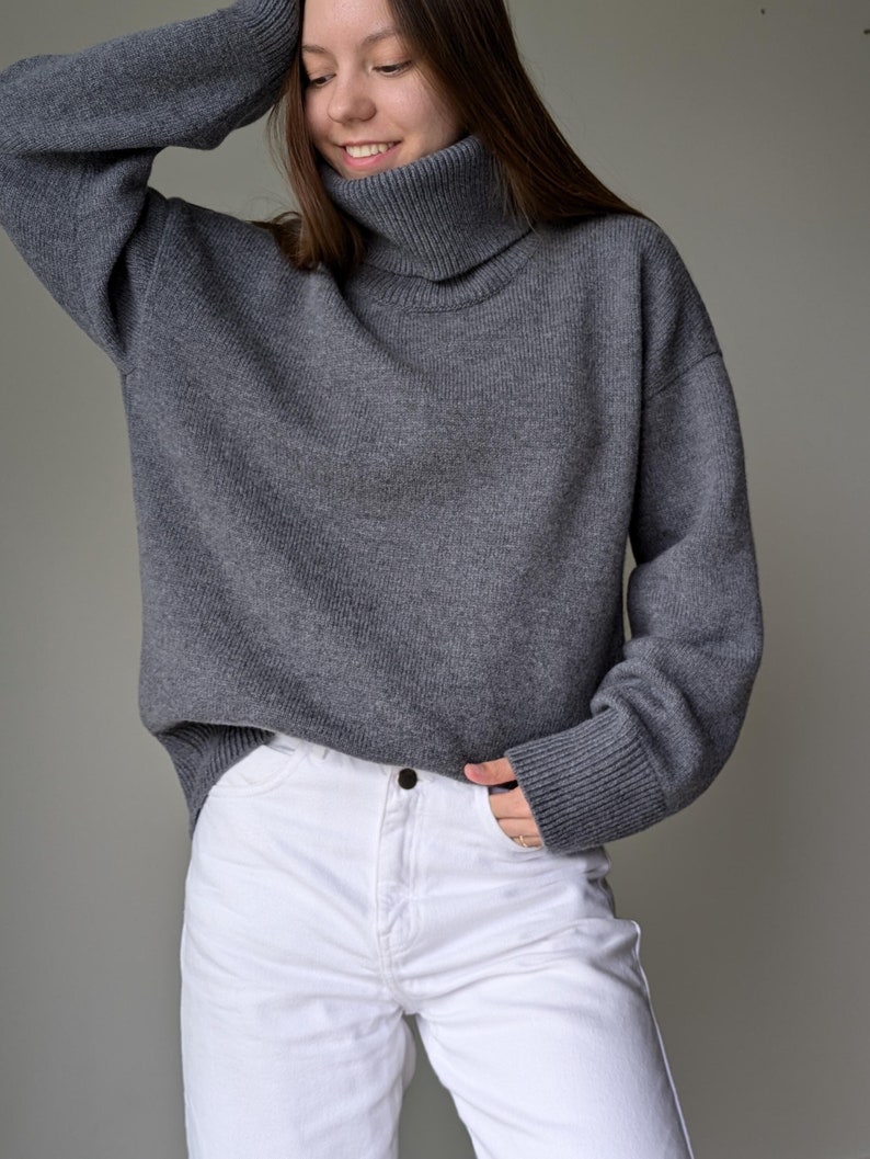 May include: A gray turtleneck sweater paired with white jeans. The sweater has a ribbed collar and cuffs. The model is smiling and has their arm raised. The jeans have a button closure and pockets.