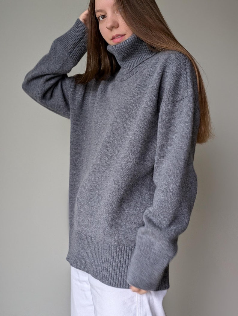 May include: A gray turtleneck sweater with ribbed cuffs and hem. The sweater is a solid color and appears to be made of a soft, knitted material. The sweater is paired with white pants.