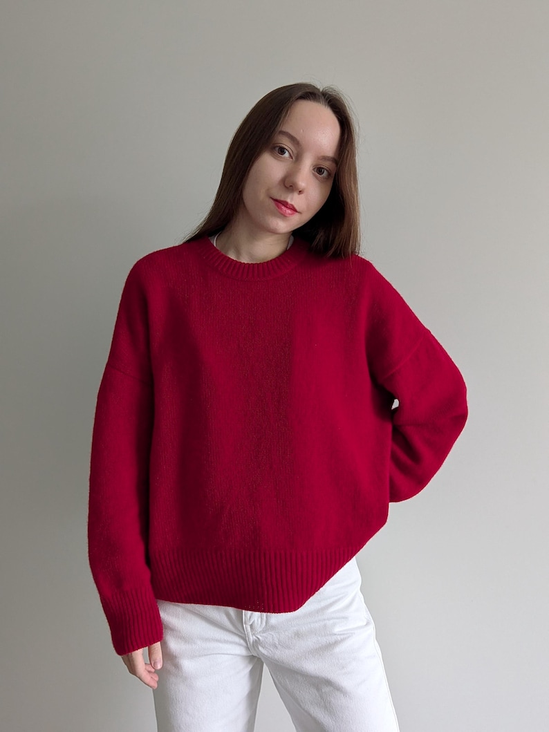 May include: A vibrant red long-sleeved sweater featuring a classic crew neckline and a ribbed knit texture at the cuffs and hem. The garment is constructed with a relaxed fit and a solid color finish, paired with plain white trousers.