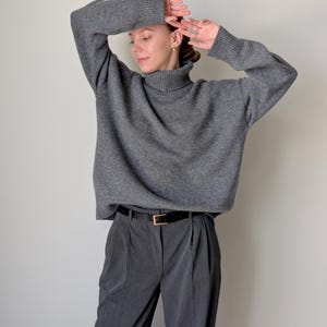 Op de afbeelding: Een antracietgrijze coltrui met lange mouwen en geribbelde boorden, gecombineerd met een donkergrijze pantalon met plooien en een zwarte riem met een goudkleurige gesp. De trui heeft een relaxte pasvorm en de broek is getailleerd.