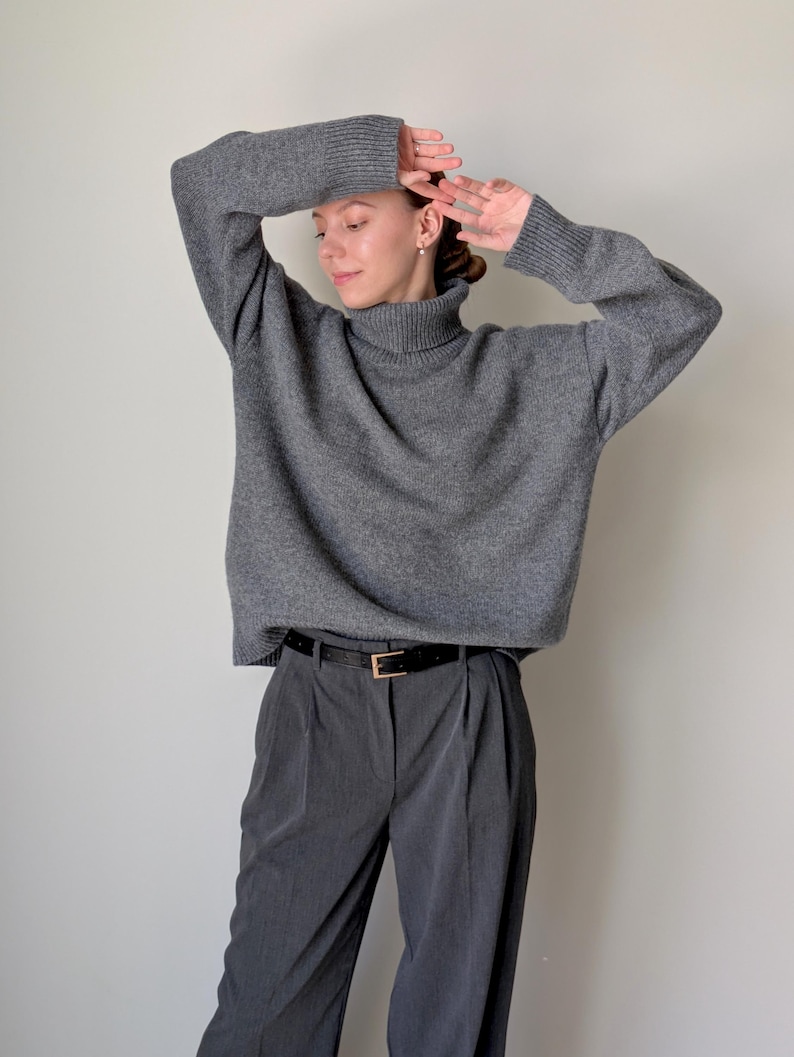 Op de afbeelding: Een antracietgrijze coltrui met lange mouwen en geribbelde boorden, gecombineerd met een donkergrijze pantalon met plooien en een zwarte riem met een goudkleurige gesp. De trui heeft een relaxte pasvorm en de broek is getailleerd.
