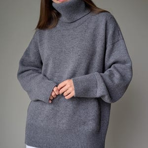 May include: A gray turtleneck sweater with ribbed cuffs and hem. The sweater has a relaxed fit and long sleeves. The model is wearing white pants. The sweater is made of a soft, knitted material.