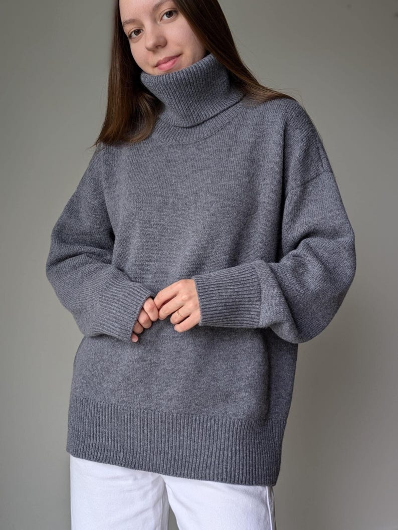 May include: A gray turtleneck sweater with ribbed cuffs and hem. The sweater has a relaxed fit and long sleeves. The model is wearing white pants. The sweater is made of a soft, knitted material.