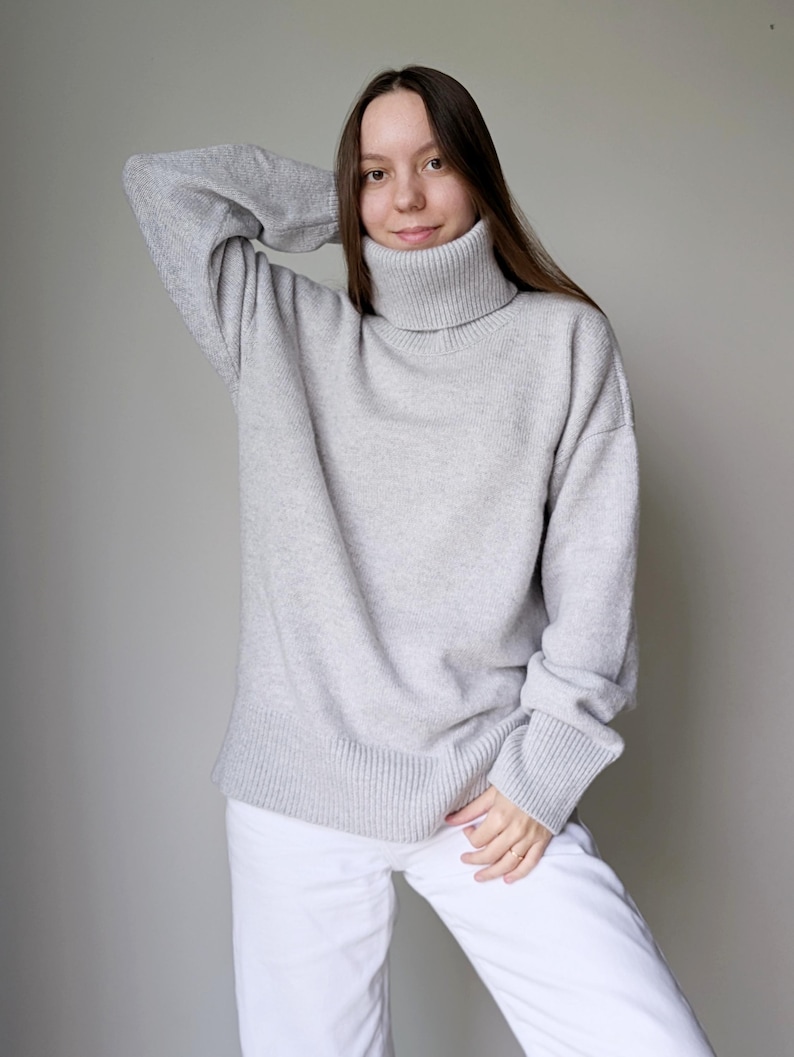 May include: A light gray turtleneck sweater with ribbed cuffs and hem. The sweater is paired with white pants. The model is wearing the sweater and pants. The sweater is a neutral color and appears to be made of a soft knit material.