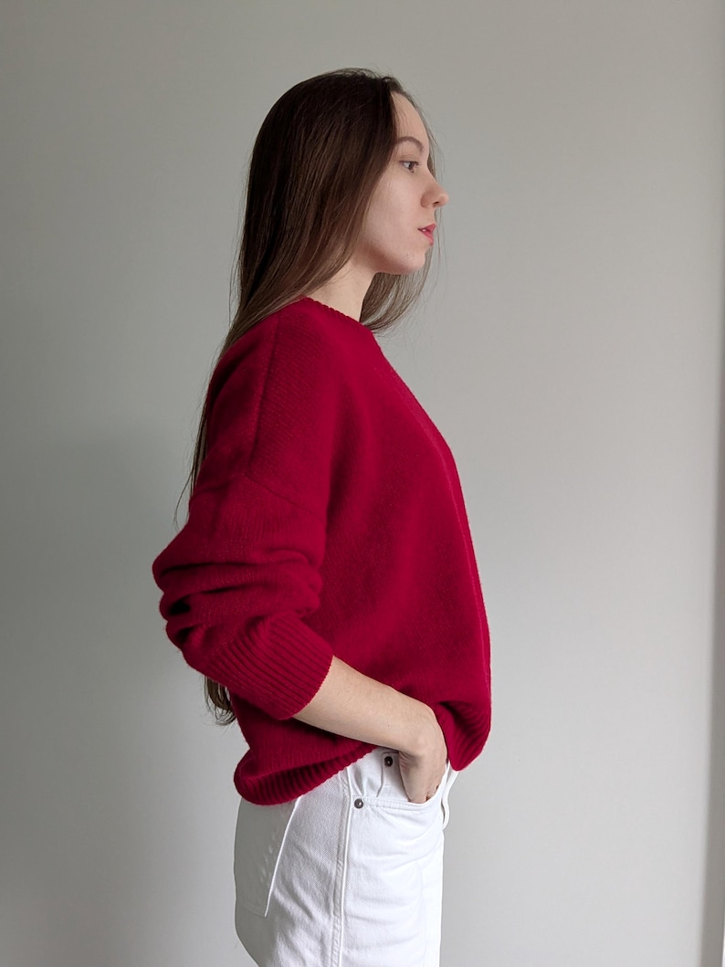 May include: A long-sleeved, solid red knit sweater featuring a crew neckline and ribbed cuffs. The sweater has a relaxed fit and is paired with white denim pants that have visible metal buttons at the waistline and front pockets.
