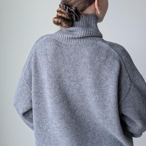 Op de afbeelding: Een grijze gebreide trui met een hoge col en lange mouwen met verlaagde schouders. Het kledingstuk is gemaakt van een gestructureerd, zacht ogend materiaal en wordt van achteren getoond, waarbij het detail van de geribbelde kraag zichtbaar is.