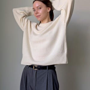 May include: A cream-colored long-sleeve knit sweater with a ribbed crew neckline, cuffs, and hem, paired with charcoal gray pleated trousers featuring a black leather belt with a gold-toned buckle, creating a neutral-toned ensemble.