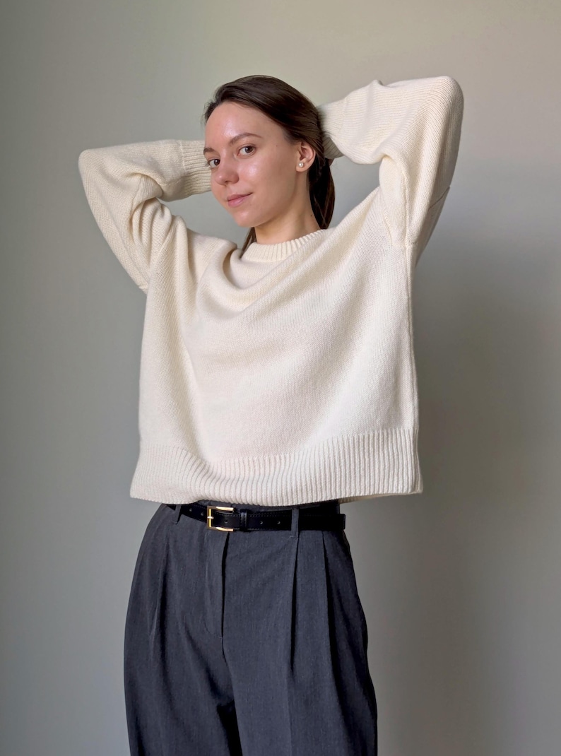 May include: A cream-colored long-sleeve knit sweater with a ribbed crew neckline, cuffs, and hem, paired with charcoal gray pleated trousers featuring a black leather belt with a gold-toned buckle, creating a neutral-toned ensemble.