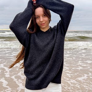 Może przedstawiać: Ciemnoszary sweter z dekoltem w serek i długimi rękawami, zestawiony z białymi spodniami. Osoba stoi na piaszczystej plaży z oceanem w tle. Niebo jest zachmurzone, a morze spokojne.