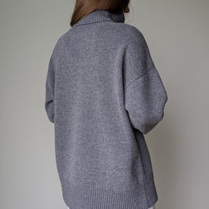 May include: A gray turtleneck sweater with a ribbed hem and cuffs. The sweater has a relaxed fit and long sleeves. The person is wearing a white garment underneath.