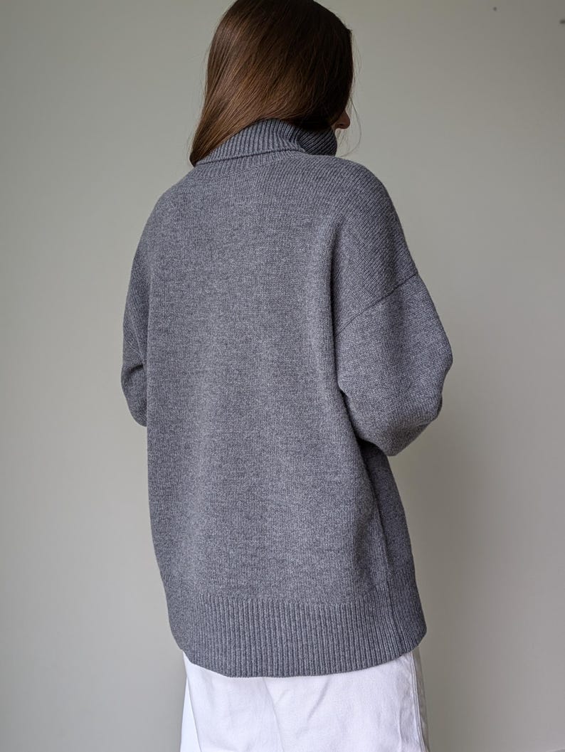 May include: A gray turtleneck sweater with a ribbed hem and cuffs. The sweater has a relaxed fit and long sleeves. The person is wearing a white garment underneath.