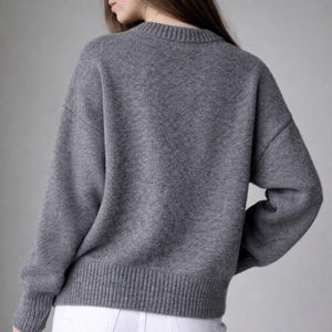 May include: A long-sleeved, charcoal gray knit sweater featuring a ribbed crew neckline, ribbed cuffs, and a ribbed hemline. The sweater has a relaxed fit with dropped shoulders and is paired with white denim pants.
