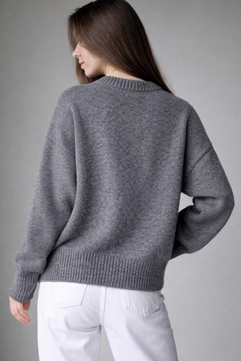 May include: A long-sleeved, charcoal gray knit sweater featuring a ribbed crew neckline, ribbed cuffs, and a ribbed hemline. The sweater has a relaxed fit with dropped shoulders and is paired with white denim pants.