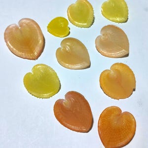 Könnte beinhalten: Eine Ansammlung von zehn herzförmigen, durchscheinenden und strukturierten Dekorationselementen in verschiedenen Gelb-, Orange- und Pfirsichtönen. Jedes Stück weist ein ausgeprägtes, von der Mitte ausgehendes Rippen- oder Adernmuster auf, das an Blätter erinnert.