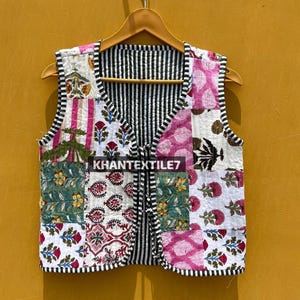 Op de afbeelding: Een gewatteerd vest met een patchwork-ontwerp, met bloemen- en geometrische patronen in roze, groen en wit. Het vest heeft een zwart-wit gestreepte binnenkant en afwerking. De tekst "KHANTEXTILE7" is zichtbaar.