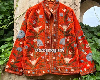 May include: An orange velvet jacket with intricate floral embroidery in white, blue, and green. The jacket features a mandarin collar and is hanging on a wooden hanger. The text "KHANTEXTILE7" is visible on the jacket.