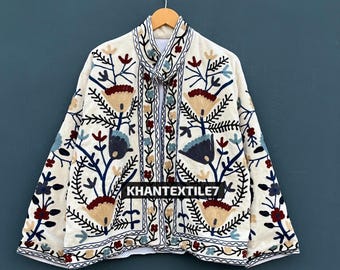 May include: Cream-colored jacket with intricate floral embroidery in shades of blue, red, and tan. The jacket features a mandarin collar and is adorned with the text "KHANTEXTILE7". The embroidery creates a detailed pattern across the jacket's surface.