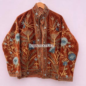 Peut inclure: Veste en velours marron avec broderies florales complexes bleues et dorées. La veste, suspendue à un cintre en bois, présente un col montant. Le texte "KHANTEXTILE7" est visible sur le devant.
