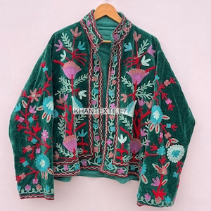 May include: A green velvet jacket with colorful floral embroidery. The jacket features a high collar and is adorned with red, blue, pink, and purple floral designs. The text "KHANTEXTILE7" is visible on the front of the jacket.