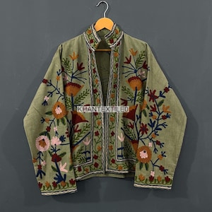 Peut inclure: Veste vert olive avec broderies florales complexes dans les tons orange, rose et bleu. La veste a un col mandarin et est suspendue à un cintre en bois. Le texte "KHANTEXTILEO" est visible sur la veste.