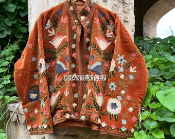 May include: Rust-colored velvet jacket with floral embroidery in white, blue, and pink. The jacket has a mandarin collar and a button-up front. The sleeves and front panels are adorned with detailed floral patterns. The text "KHANTEXTLE7" is visible.