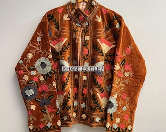 May include: A brown velvet jacket with intricate floral embroidery in shades of pink, blue, and cream. The jacket features a high collar and is hanging on a wooden hanger. The text "KHANTEXTILE7" is visible on the jacket.
