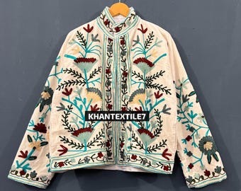 May include: Cream-colored jacket with intricate floral embroidery in teal, red, and yellow. The jacket features a mandarin collar and button-up front. The text "KHANTEXTILE7" is visible on the front. The jacket is hanging on a wooden hanger.