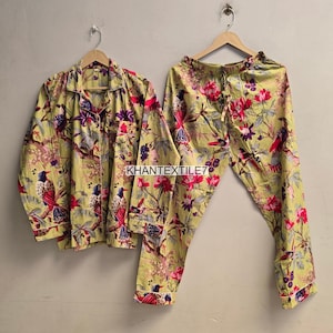 May include: A long-sleeved button-down shirt and matching pajama pants in a light yellow fabric featuring a colorful print of birds, branches, and pink flowers. The text KHANTEXTILE7 is printed in the center of the set on the light background.