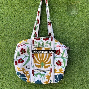 May include: White tote bag with a floral design in red, green, and yellow. The bag has long handles and a zipper closure. The text "KHANTEXTILE7" is printed on the front of the bag. The bag is sitting on a green surface.