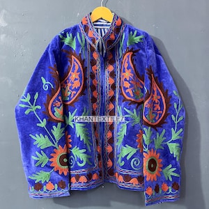 May include: A blue velvet jacket with intricate embroidery. The jacket features large paisley designs in orange and brown, with green and orange floral accents. The text "KHANTEXTILE7" is visible on the front. The jacket is hanging on a yellow hanger.