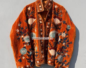 May include: An orange velvet jacket with intricate floral embroidery in white, blue, and maroon. The jacket features a collared neckline and is adorned with detailed stitching along the edges and sleeves. The overall design is a blend of textures and colors.
